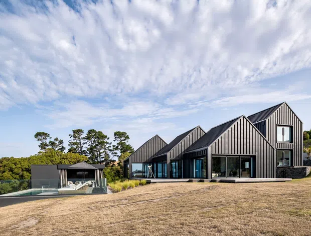 Wide exterior view of a contemporary multi-gabled residence on a sloping landscaped site. The house features repeated steep pitched gable forms, dark vertical standing-seam or ribbed cladding, extensive floor-to-ceiling glazing, and a broad terrace. A separate covered outdoor structure and swimming pool are visible to the left, with trees and distant landscape beyond.