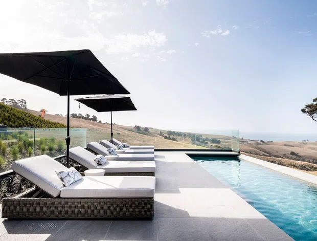 A luxurious outdoor pool terrace overlooking a dry coastal or rural landscape. Several white-cushioned woven loungers sit beneath large black umbrellas beside a long turquoise infinity-style pool, with frameless glass balustrading and pale stone paving. The scene is sunlit, spacious and resort-like.
