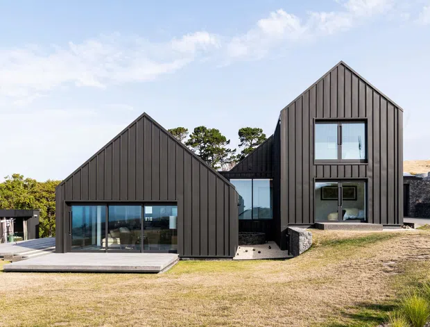 Exterior view of a contemporary multi-volume home composed of dark vertical cladding and steep gable forms. Large glazed openings connect the lower level to a broad deck, while a taller central volume includes stacked windows. The house sits in a landscaped, slightly sloping setting with exposed stone retaining elements and an additional small outbuilding visible at the side.