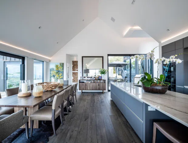 Bright, open-plan kitchen and dining area with a vaulted white ceiling, extensive glazing, dark grey full-height cabinetry, a large island with a light stone benchtop, and a timber dining table. Dark timber-look flooring runs throughout, while an indoor orchid arrangement and contemporary furnishings add warmth. The space opens visually toward an outdoor area through the glazing.