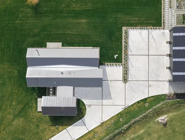 Aerial top-down view of a contemporary low-rise property set within expansive landscaped grounds. The composition shows several elongated light-grey metal-roofed building volumes, broad pale concrete driveways or paved courtyards, and a large dark rectangular roofed structure with contrasting linear skylight or roof-panel bands. Curving access paths and lawn surround the built areas.