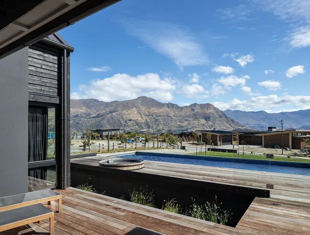 A contemporary home exterior and timber deck overlooking a swimming pool, with rugged mountains visible beyond a low-density residential setting. The facade combines dark horizontal timber cladding, smooth grey wall panels, and large glazed openings. The deck and pool area create a spacious outdoor entertaining setting.