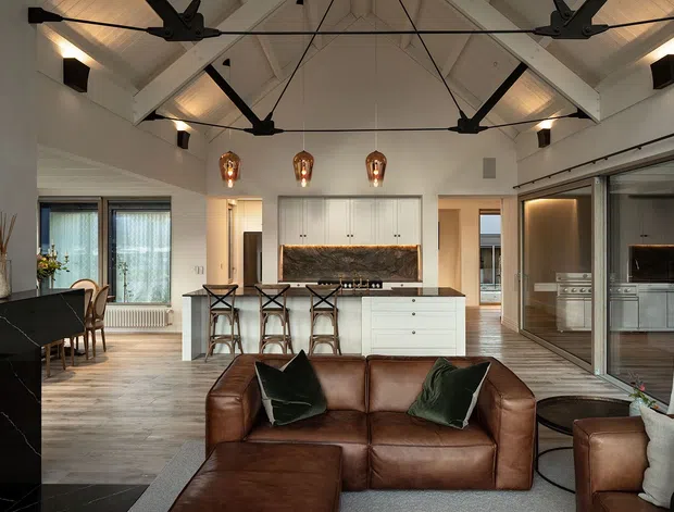 Open-plan contemporary living, dining and kitchen interior with a dramatic pitched, exposed-beam ceiling. The room features a white kitchen with central island and bar seating, dark stone surfaces, timber flooring, extensive sliding glass doors, black ceiling fans and warm pendant lighting. A brown leather sectional sofa occupies the foreground, with dining furniture and a secondary kitchen or utility zone visible beyond the glass partition.