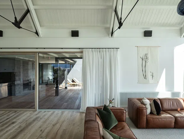 Bright contemporary living room with a high white paneled ceiling, exposed dark structural bracing, warm timber-look flooring, and large glazed sliding doors opening to a timber deck. Brown leather sectional sofas, neutral cushions, white curtains, and simple botanical wall art create a refined, airy interior.