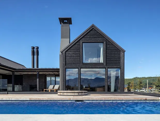 Exterior view of a contemporary dark-clad gable-roof house beside a swimming pool. The facade features vertical glazing, black timber-style cladding, a prominent chimney, and an attached low-pitched wing with covered outdoor living space. Mountainous terrain is visible in the distance.