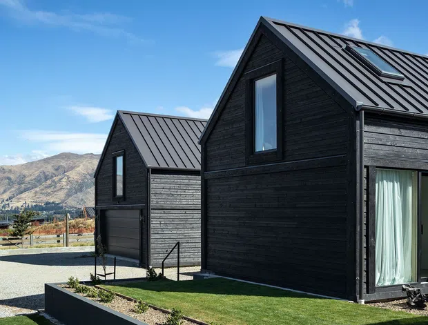 Exterior view of a contemporary dark-clad gabled house composed of connected volumes. The building features black stained horizontal timber cladding, standing-seam metal gable roofs, tall glazed openings, and a roof skylight. A landscaped lawn and gravel garden border the foreground, with rugged mountains visible in the distance.
