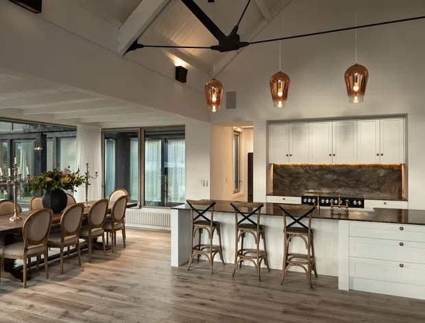 Open-plan kitchen and dining area with white cabinetry, a dark stone feature splashback, central island with bar seating, timber flooring, and a large dining table beside expansive glazed doors. The room features a vaulted ceiling with exposed painted beams, a dark ceiling fan, and three warm pendant lights.