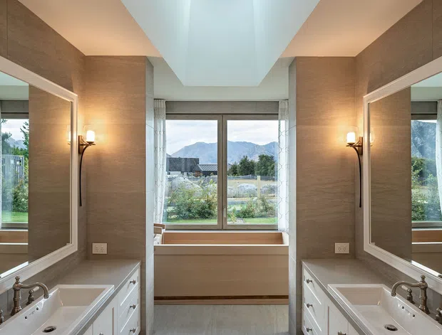 Bright symmetrical bathroom with twin white vanity units, large framed mirrors, warm textured taupe walls, a central window seat beneath mountain-view glazing, and a skylight above. Wall sconces provide warm accent lighting, while pale flooring and cabinetry create a refined, spacious feel.