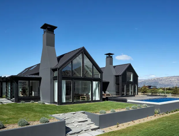 Exterior view of a contemporary multi-gabled house with dark metal roofing, extensive floor-to-ceiling glazing, tall rendered chimneys, and dark framed window systems. The landscaped grounds include clipped lawns, low concrete retaining walls, stone steps, and a rectangular swimming pool. Mountains are visible in the distant background under a clear blue sky.