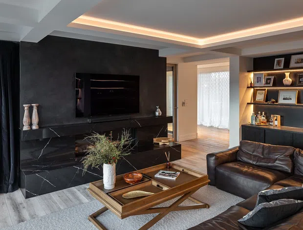 A sophisticated contemporary living room with a black marble feature fireplace and television wall, integrated illuminated display shelving and bar cabinetry, dark leather sectional seating, and a timber coffee table. Recessed cove lighting, layered neutral finishes, and large curtains create a refined, moody atmosphere.