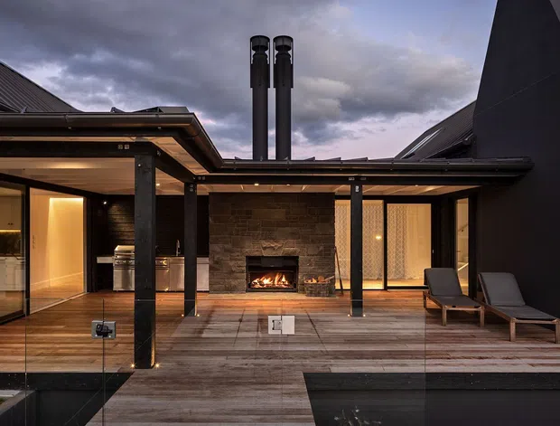 A dramatic contemporary courtyard terrace viewed from outside, with extensive dark-framed glazing, timber decking, a central stone-clad outdoor fireplace, black structural posts, outdoor lounge chairs, and tall chimney flues beneath a moody overcast sky. Warm interior lighting from the adjoining rooms contrasts with the dark exterior finishes.
