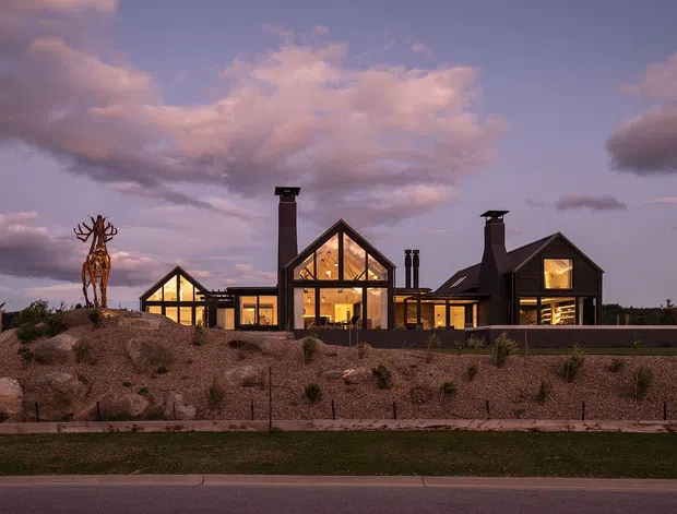 Exterior dusk view of a sprawling contemporary residential property with multiple steep gabled volumes, dark cladding, prominent floor-to-roof glazing, and illuminated interiors. The house sits within a landscaped, rocky site with sparse planting, low retaining walls, and a sculptural tree-like installation.