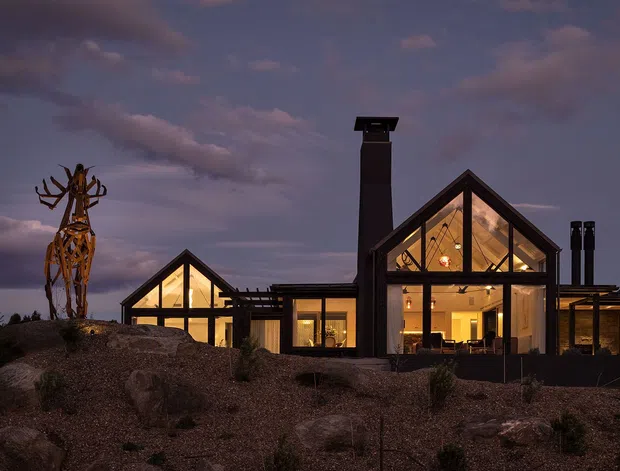 Exterior dusk view of a contemporary multi-gabled house with extensive floor-to-ceiling glazing, dark structural framing, prominent chimneys, and warm interior lighting. The building sits in a sparse landscaped setting with gravel, rocks, and sculptural planting.