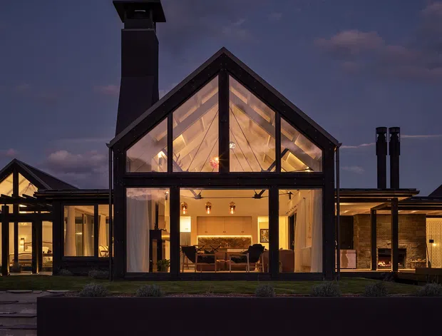 Twilight exterior view of a contemporary gable-roofed house with a dramatic double-height glazed central living space. Dark structural framing and cladding contrast with the warm illuminated interior, while adjoining low-slung wings, a tall chimney, and outdoor fireplace elements are visible.