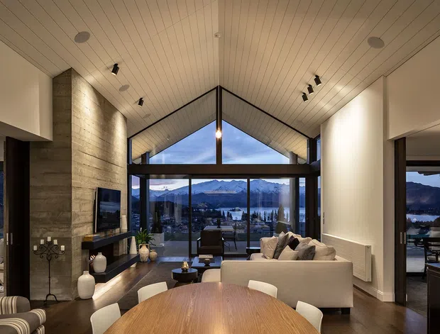 A refined open-plan living and dining area with a dramatic vaulted timber-lined ceiling and full-height glazing overlooking a mountainous lakeside landscape. The room features a large round timber dining table, contemporary upholstered seating, a stone fireplace wall with built-in television, dark flooring, and warm layered lighting.