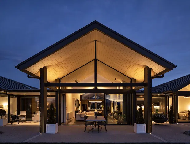 Dusk exterior view of a low-rise contemporary home centered on a dramatic gabled pavilion. The pavilion has a deep projecting roof, warm timber-lined soffit, dark structural framing, extensive floor-to-ceiling glazing, and an illuminated open-plan interior visible beyond. Symmetrical single-storey wings extend to either side, with outdoor seating and large planters in the foreground.
