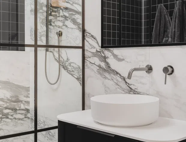 Contemporary bathroom featuring dramatic white marble-look wall and floor slabs with dark veining, black square-tile accents, black-framed glazing or mirrors, a frameless glass shower enclosure with wall-mounted shower fittings, and a white vessel basin on a dark floating vanity. Brushed metal tapware adds a restrained metallic detail.