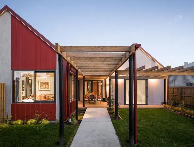 Exterior view of a contemporary residential property with a red vertical-clad gabled volume, pale rendered walls, timber pergola-covered walkway, concrete path, lawn, and timber fencing. Glazed doors and windows reveal a warm interior dining or living area, while a second gabled volume and low roof forms extend behind the courtyard-like approach.