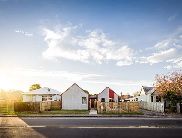 Street-facing view of a low-rise residential neighbourhood with several detached homes, front fences, driveways and lawns under a bright sky. The central home has a simple white gabled facade with a contrasting red side wall, while neighbouring houses show pitched roofs and weatherboard-style exteriors. The image is an exterior streetscape photograph.