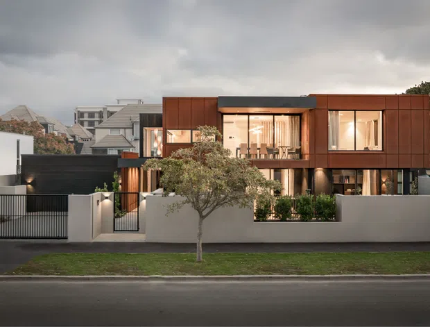 Street-facing view of a contemporary multi-level residence with a rectilinear form, dark metal roof, and warm rust-brown metal or timber-look cladding to the upper facade. Large glazed openings reveal a warmly lit interior, while a white rendered boundary wall, black metal gates, and landscaped planting define the front setback. The surrounding suburban streetscape and overcast sky are visible.