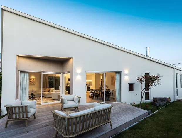 Exterior view of a contemporary single-storey residence with a simple white rendered facade and dramatically sloping roofline. Large glazed openings connect the interior living and dining spaces to a timber deck furnished with outdoor seating. The image also shows a narrow paved side path, lawn, and a small landscaped courtyard area.