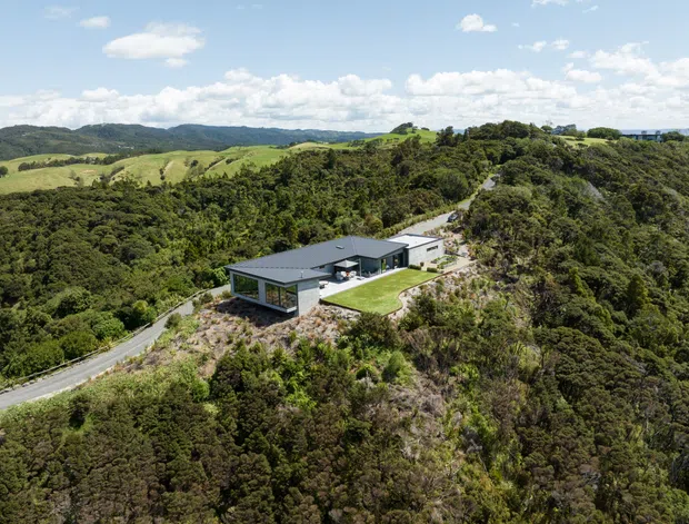 Aerial view of a low, contemporary single-storey house with dark flat or low-pitched roofs, extensive glazing, and a small lawn terrace. The home is positioned on a steep, heavily vegetated ridge with a winding access road, overlooking rolling rural hills.