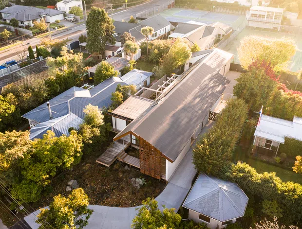 Aerial view of a leafy suburban residential property featuring a long, narrow building with a pitched corrugated roof, timber-clad walls and connected outdoor paths or decks. Several smaller residential structures and landscaped gardens surround the main building, with a road and neighbouring homes visible beyond.