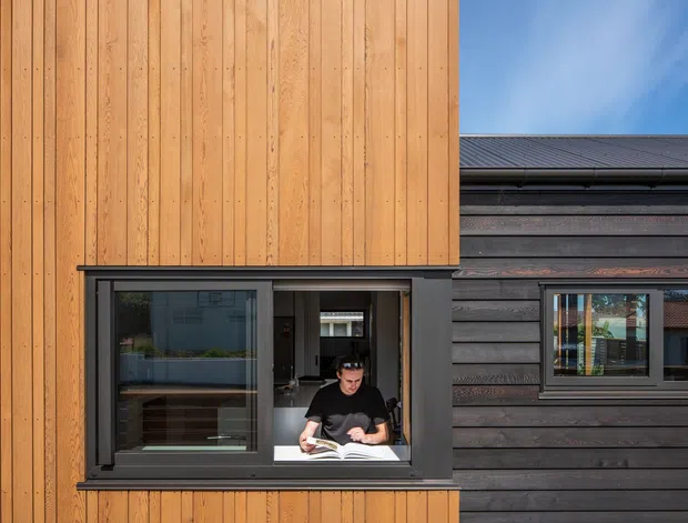 Contemporary building facade clad in warm vertical timber boards and dark horizontal weatherboards. A projecting or recessed window opening forms a small study or reading nook, with a person seated at a desk inside. The composition highlights contrasting timber cladding textures, dark window frames, and a low-pitched roof edge against a blue sky.
