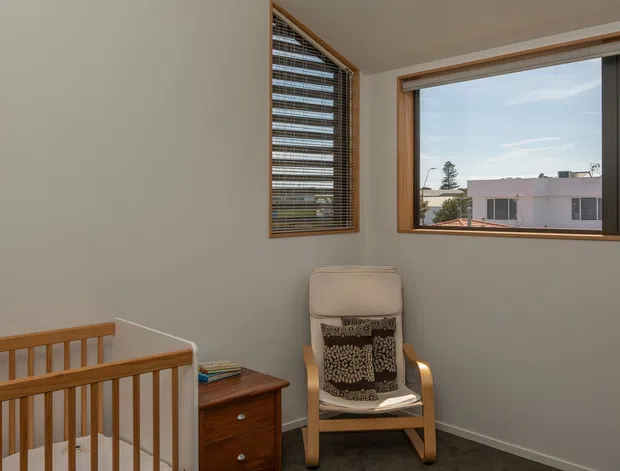 A bright, compact nursery with a timber cot, wooden chest of drawers, upholstered rocking chair, and patterned cushion. The room has soft off-white walls, dark grey carpet, timber-framed windows with blinds, and warm natural light. A residential neighbourhood is visible through the windows.