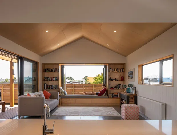 Bright open-plan living room with a pitched timber-lined ceiling, white vertical wall panelling, built-in shelving and window-seat joinery, expansive glazing, and a kitchen benchtop in the foreground. The room opens toward a deck with coastal views and features a neutral palette with warm timber accents.