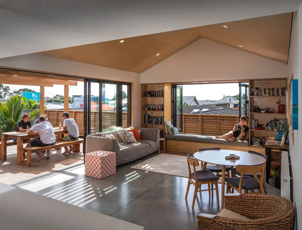 Bright, open-plan living and dining area with a distinctive angular timber-lined ceiling, polished concrete floor, extensive glazing, built-in shelving, and an adjoining timber deck. A sofa, round dining table, window seat, and casual outdoor dining area create a relaxed contemporary family space.