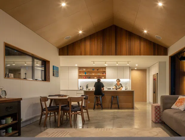 Warm, open-plan contemporary living and dining area with a vaulted timber-lined ceiling. The room combines a timber dining table and chairs, a compact kitchen with a central island and stools, built-in cabinetry, polished concrete flooring, and a comfortable sofa seating area. Vertical timber panelling forms a feature wall beneath the gable, while integrated downlights create a welcoming evening ambience.