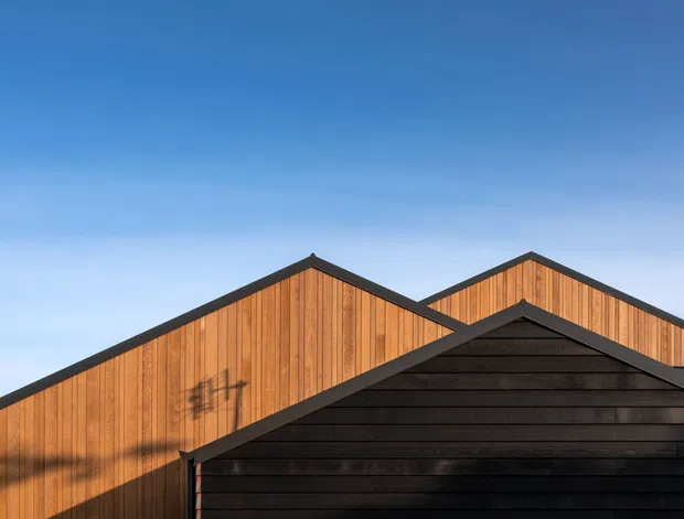 Close-up exterior view of overlapping gabled building forms with vertical warm timber cladding and a dark horizontal-clad foreground gable. Strong geometric rooflines are set against a clear blue sky.
