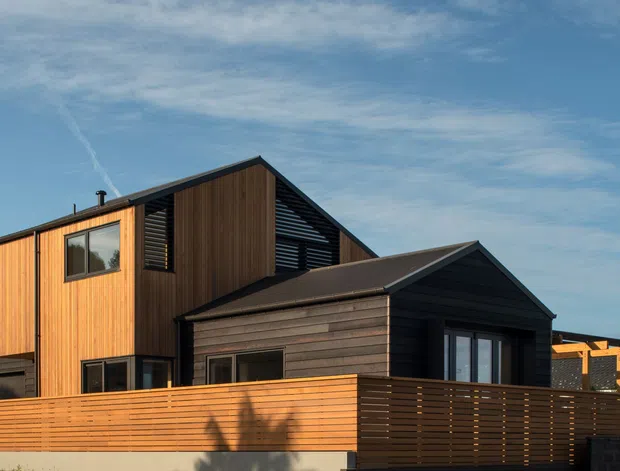 Exterior view of a contemporary multi-level house with a steep gabled roof, contrasting vertical timber cladding on the upper volume and dark horizontal cladding on the lower front volume. A horizontal timber privacy fence and dark-framed windows define the foreground facade.