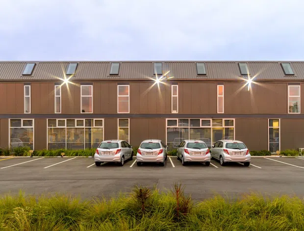 A contemporary multi-storey residential or mixed-use building viewed from across a paved parking area. The long facade features dark brown vertical cladding, a shallow pitched metal roof with skylights, rows of windows, and recessed balconies with timber-toned soffits. Several parked cars and landscaped grasses occupy the foreground.