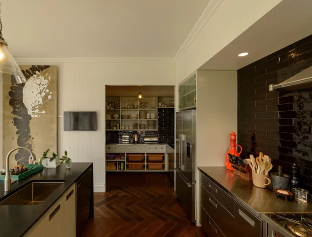 A warm, refined contemporary kitchen viewed toward a compact butler's pantry or scullery. The main kitchen has dark benchtops, dark glossy subway-tile splashbacks, extensive dark cabinetry, a contrasting light island with an integrated sink, stainless-steel appliances, and a rich dark timber herringbone floor. White vertical wall panelling, glass-fronted storage, pendant lighting, and open pantry shelving complete the space.
