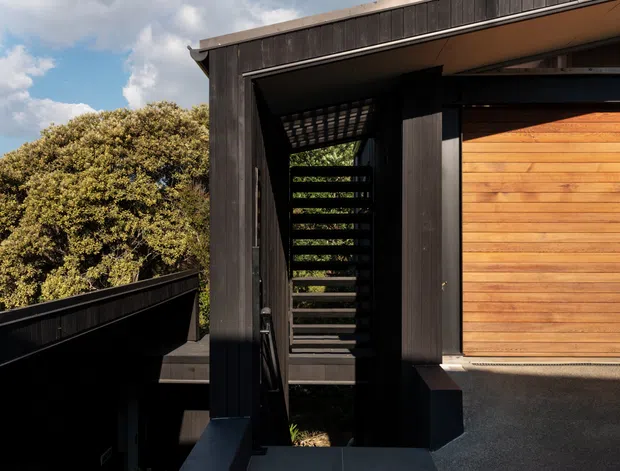 Exterior view of a contemporary dark-clad building entrance or circulation zone, with a black timber-clad facade, covered stairway with horizontal slatted screening, warm timber garage or entry door, and dark paved surfaces. Dense vegetation and sky are visible around the structure.