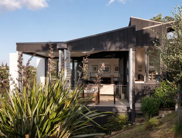 Exterior view of a contemporary single-storey house with dark vertical timber cladding, a low-pitched roof, and an open covered terrace or entry space. Large glazed openings reveal a light-filled interior living area with framed artwork and built-in furnishings. Native-style planting and landscaped garden surround the home.