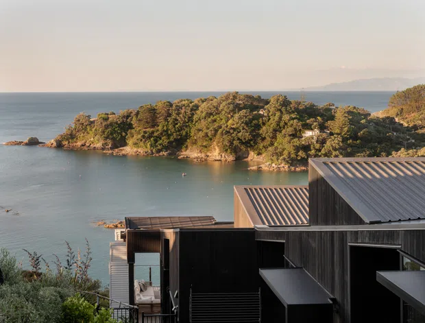Exterior view of a contemporary dark-clad, multi-level coastal house positioned above a sheltered bay. The home has overlapping pitched metal roofs, strong angular volumes, extensive glazing, and timber-toned dark vertical cladding. A wooded peninsula, calm water, and distant hills form the surrounding setting.