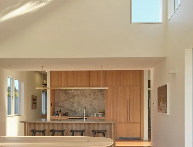 Bright, double-height open-plan kitchen and dining area with warm timber cabinetry, a large stone feature splashback, island breakfast bar with stools, and a long timber dining table with benches. Tall windows and clerestory glazing fill the space with sunlight, while polished timber flooring and neutral walls create a warm, contemporary interior.
