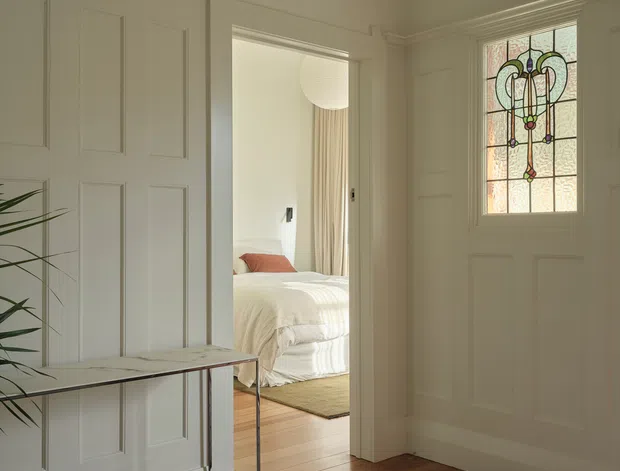 A bright, refined residential entryway with white paneled walls and doors, detailed cornices, a timber floor, and a large patterned rug. A slim console table sits against the panelled wall, while a stained-glass front door provides a decorative focal point. Through the open doorway, a softly styled bedroom with a bed, curtains, and timber flooring is visible.