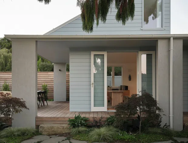Front exterior view of a light grey weatherboard house with a covered entry porch, open glass door, pale timber decking, simple vertical columns, and a landscaped garden. Irregular large stone paving forms the foreground path, while mature trees partially frame the facade. The image also reveals a glimpse of the kitchen and dining area through the open doorway.