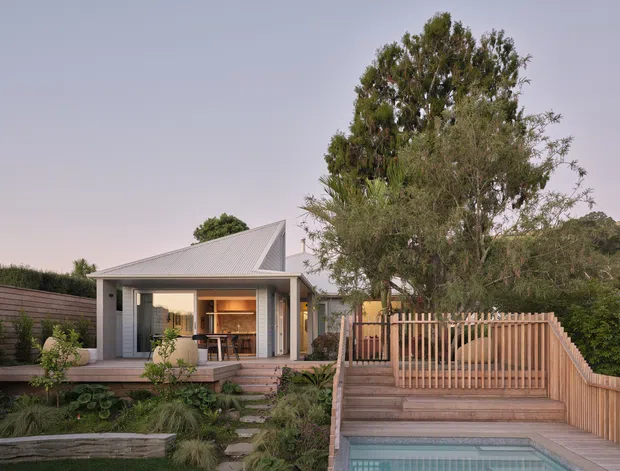 A compact contemporary house viewed from the landscaped garden, featuring a steep asymmetrical pitched metal roof, extensive glazed openings, a timber deck and steps, and a raised timber pool terrace with a pale blue swimming pool. Native-style planting, timber fencing, and warm interior lighting create a refined indoor-outdoor connection.