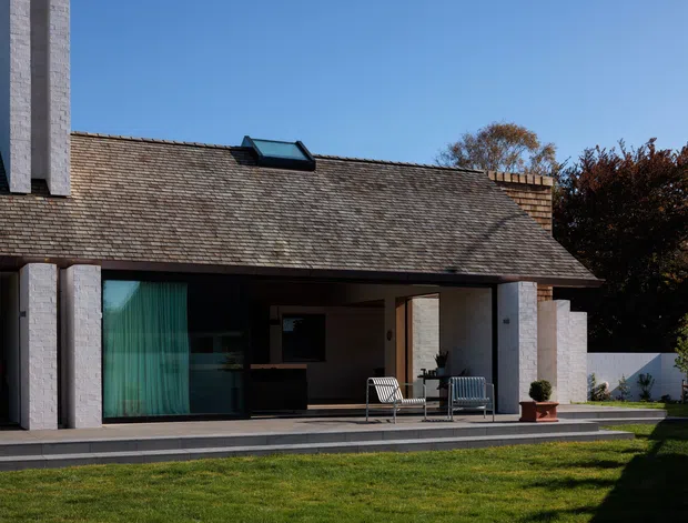 Exterior view of a low-rise modern residence with a broad pitched shingle roof, prominent pale masonry chimney and structural piers, dark-framed glazing, and a covered patio with outdoor seating. The house opens onto a paved terrace and lawn, with a small roof skylight and enclosed courtyard-like garden edge visible.