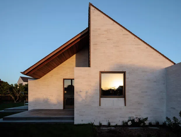 Exterior architectural photograph of a compact contemporary house with intersecting pitched roof forms, pale masonry walls, deep timber-lined eaves, dark-framed glazing, and a recessed entry terrace. The image is taken at dusk or in warm low-angle light, with a clear blue sky and landscaped garden surrounding the building.