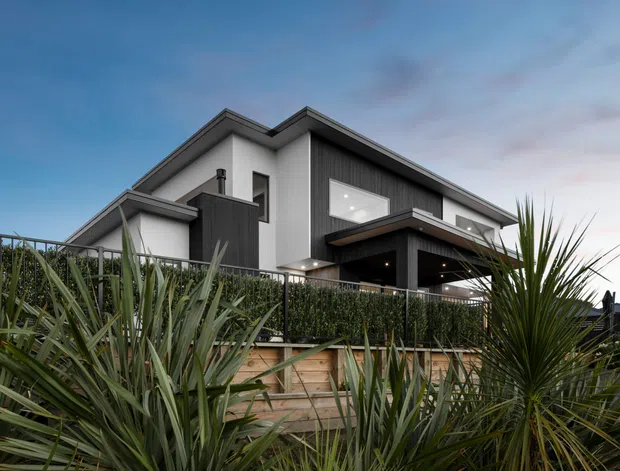 Exterior view of a contemporary multi-level home at dusk, featuring crisp white rendered walls, dark vertical cladding, angular intersecting roof planes, expansive windows, and a covered upper terrace. The house is partially screened by dense architectural planting, hedges, and timber retaining elements.