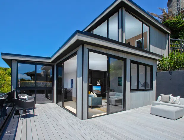 Contemporary multi-level house exterior with dark-framed floor-to-ceiling glazing, pale vertical cladding, and a steep gabled roof form. The home opens onto a broad light timber deck furnished with outdoor seating and a dining area, with interior living spaces visible through the glazing.