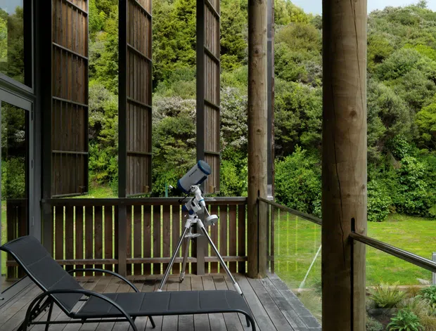 A covered timber outdoor deck or veranda overlooking a lush landscaped garden. The space features weathered timber decking, large round timber posts, vertical timber privacy screens, a dark metal reclining lounge chair, a telescope on a tripod, and a simple timber bench. Glazed doors or windows are visible at the left edge, while the open-sided structure creates a sheltered connection to the garden.