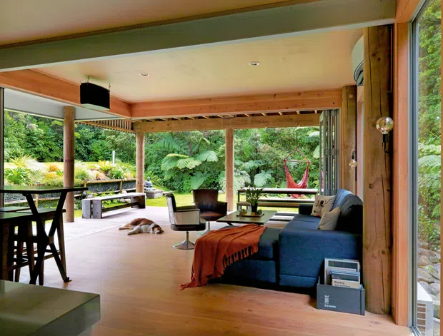 A warm, open-plan living room with extensive glazing opening onto lush garden surroundings. The space features timber flooring, exposed timber posts and beams, a light timber ceiling, a blue sectional sofa, lounge chairs, and a dining area at the left. Large sliding glass doors connect the interior to a covered outdoor terrace, creating an airy indoor-outdoor setting.