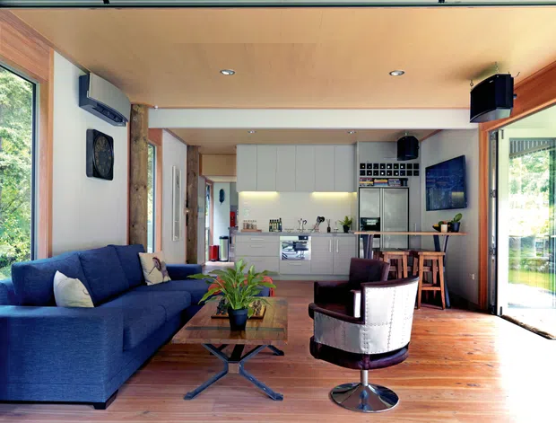 Open-plan living and kitchen interior with a deep blue sectional sofa, timber flooring and ceiling, white built-in kitchen cabinetry, a compact island, dining stools, and large glazed openings connecting to greenery outside. The room includes a small seating area and visible audiovisual equipment, creating a bright, relaxed contemporary space.